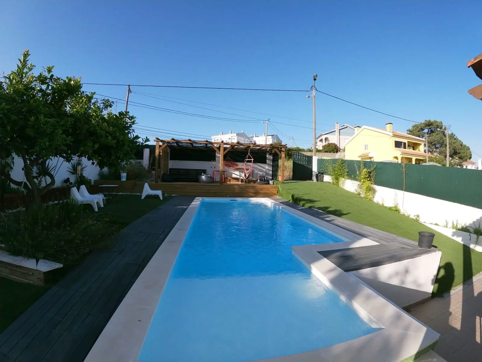 Piscina comprida entre o relvado e o deck, com a pérgula ao fundo